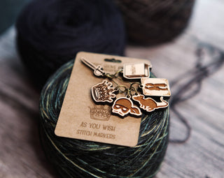 A set of Princess Bride Knitting Stitch Markers—featuring charming metal designs such as a crown, sword, skull, and whale—rests atop a ball of multicolored yarn. The card reads AS YOU WISH STITCH MARKERS.