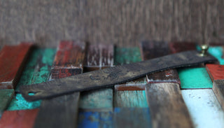 The Bee and Moon Leather Ruler Cuff features a weathered metal ruler with a brass fastener, upcycled into a stylish bracelet resting on colorful reclaimed wood in shades of red, green, blue, and brown.