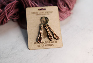 A set of three bronze-colored 🧹 Witch’s Broom Stitch Markers 🧹 shaped like potion spoons, attached to a kraft paper card, perfect for knitting or crochet projects, with purple yarn in the background.