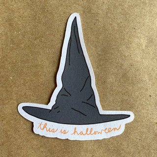 The This is halloween witch hat sticker features a tall black hat with this is halloween in orange cursive. Eco-friendly and perfect for Halloween decor, its shown on a brown textured surface.