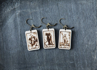 Three rectangular wooden stitch markers etched with tarot cards—The Hermit, The Magician, and Strength—rest on a textured dark gray background. Each features a metal hook and can also be used as unique knitting earrings.