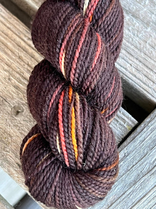 A skein from the KPM and KPPPM Variegated Color Collection rests on a weathered wooden surface, featuring dark brown shades with subtle orange, yellow, and red streaks—perfectly soft and twisted for hand knitting projects.