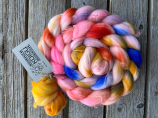 The Koigu Merino Roving Braid features a vibrant mix of pink, orange, blue, gold, and cream merino wool coiled on rustic wood. It includes a matching spinning fiber sample and a Koigu tag attached to the braid.