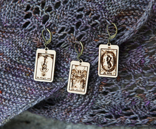 Three Tarot Card Stitch Markers—The Fool, Death & Temperance—rest on a purple and gray knitted fabric, each with an intricate black illustration on a light background. Unique crochet accessories for mystical flair!.