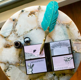 A round marble table holds an ink bottle, a turquoise feather quill with gold handle, and an open Edgar Allan Poe Sticky Notes booklet filled with sketches and quotes like “NEVERMORE.”.