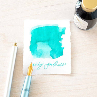 A square white card with turquoise watercolor and handwritten "inky goodness," surrounded by a fountain pen, pen cap, and a bottle of Bottled Ink, displayed on a light wooden surface.