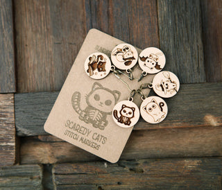 The 🐾 Scaredy Cats Stitch Marker Set 🐾 features six maple wood stitch markers with engraved cat faces on a metal ring—perfect Halloween knitting accessories, complete with a cute cat illustration and shown on a rustic wooden surface.