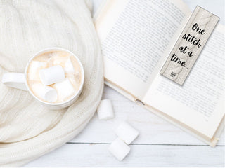 A cup of hot chocolate with marshmallows sits on a white knit blanket next to an open book. Nearby is a Printable Bookmark for Knitters that reads "One stitch at a time." Extra marshmallows rest on the white wooden surface.