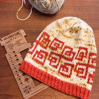 The Lonely Hearts Club KAL | 2026 Knitting Pattern hat, featuring red and yellow geometric designs, rests on a wooden table beside a yarn gauge tool and a ball of variegated yarn.