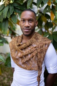 Black man in a white tee shirt wearing a brown and rust-toned shawl in front of a background of greenery