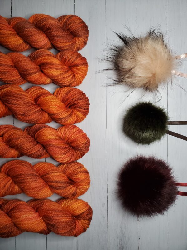 6 skeins of indie dyed yarn in deep brilliant orange with purple lined up vertically on the left, with three ikigai pom poms on the right