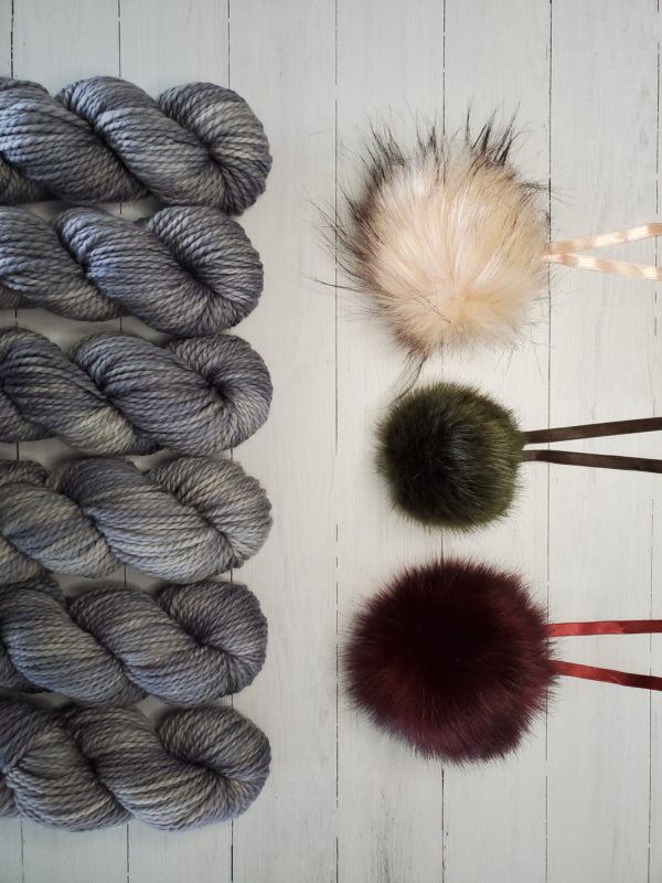 6 skeins of indie dyed yarn in grey lined up vertically on the left, with three ikigai pom poms on the right