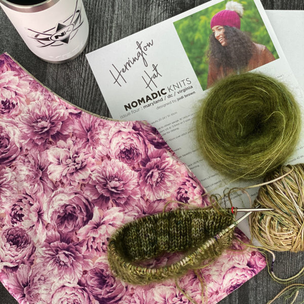 work in progress with travel mug, project bag, green mohair, a hat about 1" into the brim on the needles; herrington hat knitting pattern topped with a pom pom by jodi brown of grocery girls knit, found in Nomadic Knits knitting magazine subscription