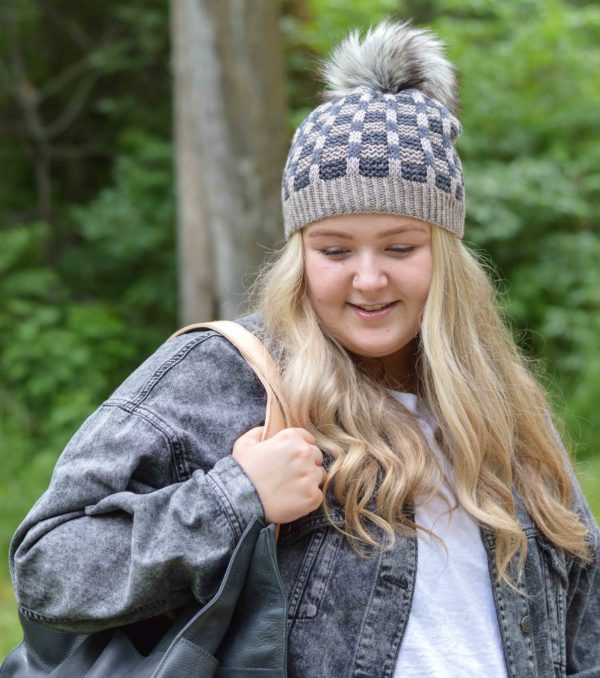 Jodi Brown's blond haired daughter modeling Higgins Lake hat knitting pattern; a blue and grey mosaic pattern, topped with a pom pom; shown in colorways from indie yarn dyer ajhc; available in Nomadic Knits knitting magazine subscription