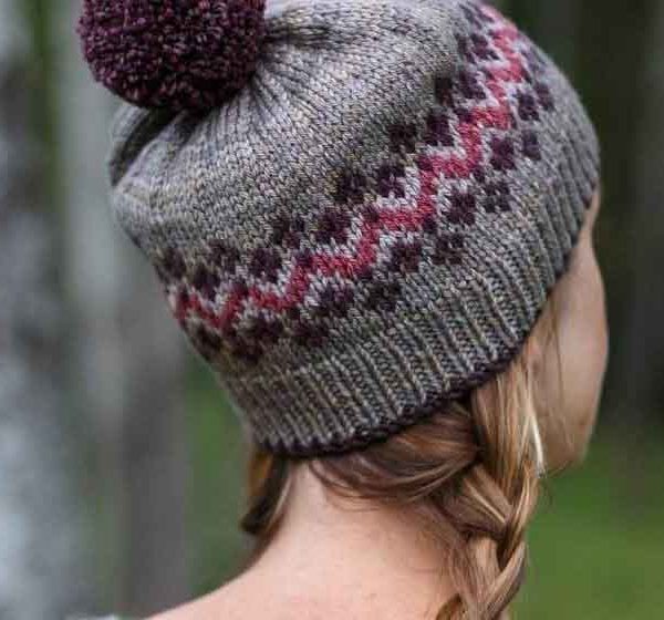 White woman with a blond braid wearing a grey hat with pink and purple Fair Isle and a purple pom pom