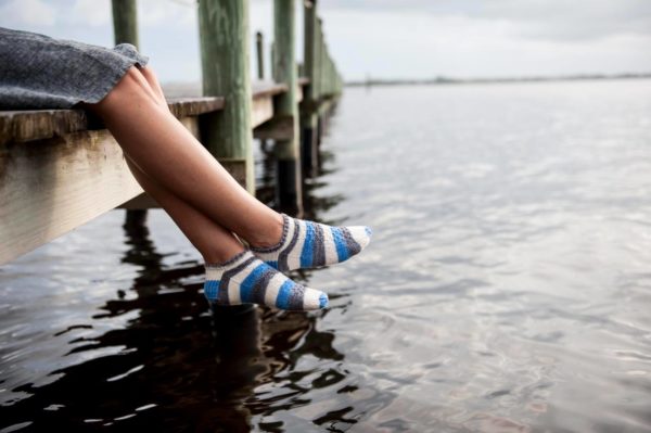 A woman is sitting on a dock over the water; only her legs and feet are visible and she is wearing short grey, blue and white striped socks. The sock knitting pattern can be found in Nomadic Knits creative knitting magazine. The indie dyed yarn used is from indie yarn dyer String Theory colorworks