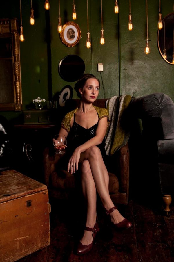Woman holding drink in Green Room of 205 Dry restaurant; there are Edison lights hanging from the ceiling in a darkened room. She is wearing a little black dress, black heels, and a sparkly pale green mohair and silk shrug with beads. The yarn is from indie yarn dyer Artyarns, and the knitting pattern can be found in Nomadic knits creative knitting magazine.