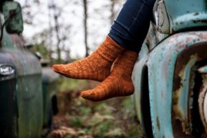 knees-down view of a woman sitting on an old truck; she is wearing burnt orange textured socks over skinny jeans. The knitting pattern is Hell Gate by Deborah Breland, knit using indie dyed yarn from The Periwinkle Sheep