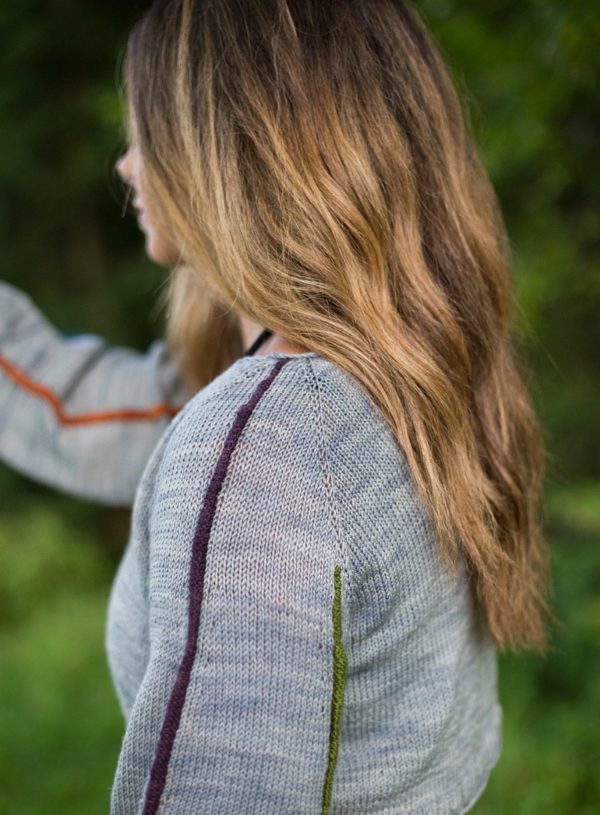 view of a woman from the side; she has flowing hair and is wearing a grey pullover cropped sweater with purple and orange skinny stripes down the sleeves. The sweater is a knitting pattern by Melissa Kemmerer, knit in Round Mountain Fibers, Vermont wool