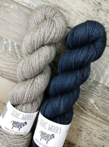2 twisted hanks of indie dyed yarn from indie yarn dyer AJHC; the skein on the left is a beautiful gray/ grey and the one on the right is a deep navy. Both colorways were used in the Higgins Lake hat knitting pattern by Jodi Brown of Grocery Girls knit, and paired with a pom pom; found in Nomadic Knits knitting magazine subscription