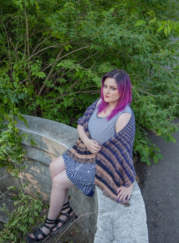 beautiful white woman with hot pink hair wearing a purple and sand striped shawl, sitting on a rock wall