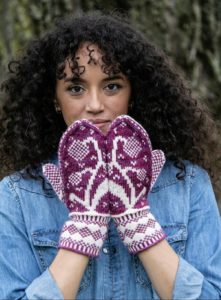 Kayla Maressa wearing a denim shirt and modeling maroon and pale pink mittens with a dandelion motif on the top side of the hands. It is the dandelion mittens knitting pattern by tanis gray knit in yarn from indie yarn dyer little fox yarn