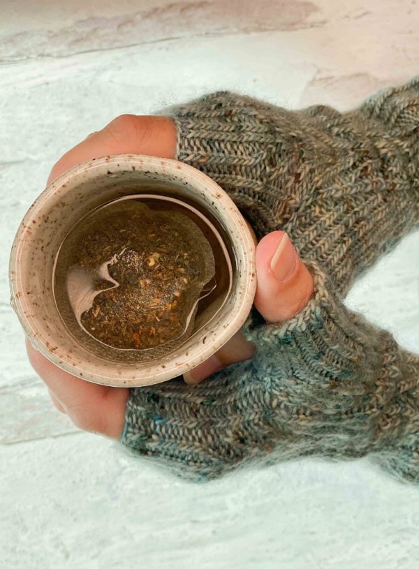 Two hands clad in greyish-green knitted mitts with cables, holding a mug filled with tea. The knitting pattern is by Kate Atherley and can be found in Nomadic Knits creative knitting magazine