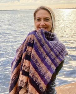 Blond woman in front of a river, wrapped in a tan and purple striped shawl. The knitting pattern is by Rizzaknits, using indie dyed yarn from Twisted Fiber art; found in Nomadic Knits creative knitting magazine