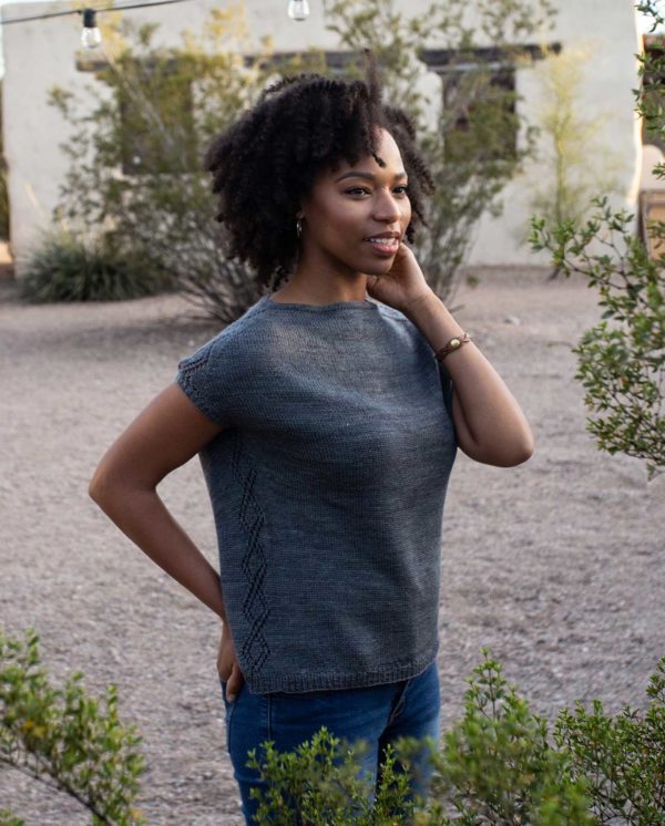 Light skinned Black woman wearing a knitted tee in turquoise, against a backdrop of desert foliage; Ocotillo knitting pattern in Nomadic Knits creative knitting magazine
